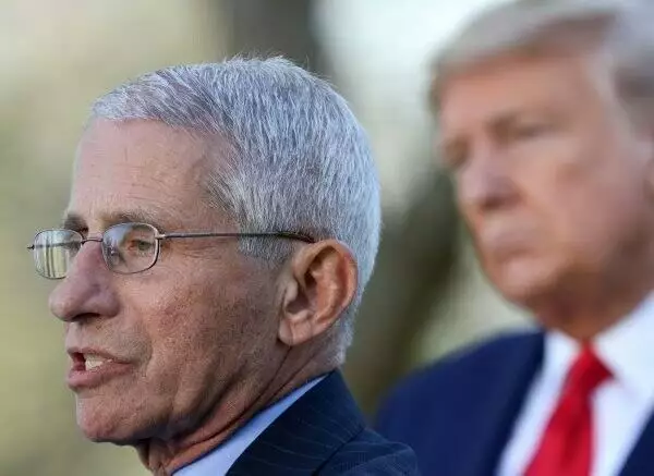 fauci in a suit and light blue shirt speaking outside with trump listening in the background