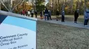 an absentee ballot drop box is in the foreground in georgia, while voters stand in line in the background.